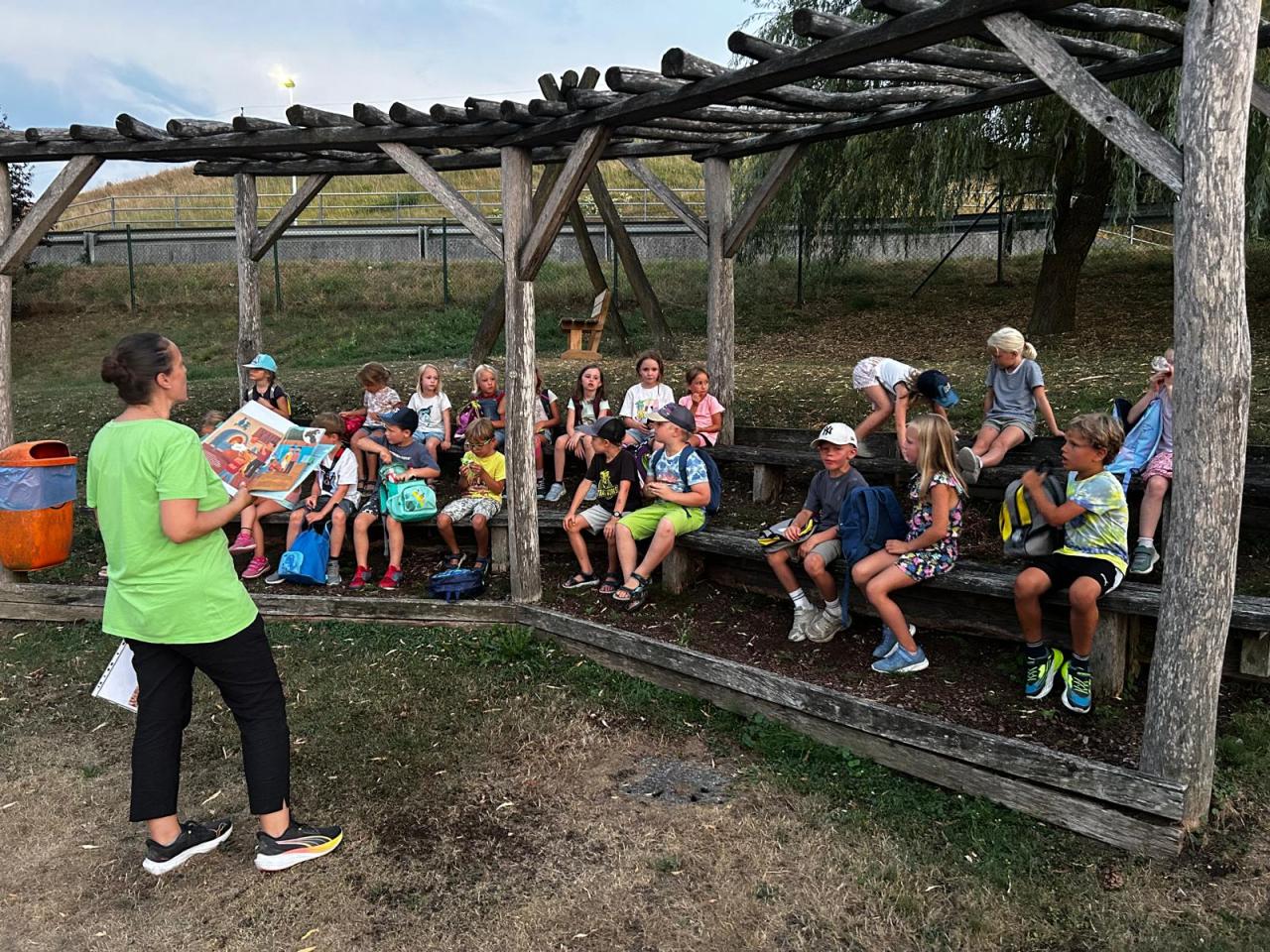 Kinder sitzen im Freien und hören einer Frau zu